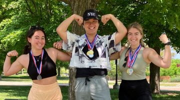 3 college students stand on an awards podium with medals around their necks as they flex.