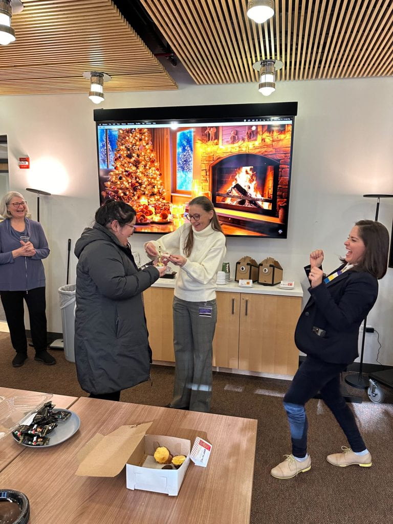 A woman in a jacket receives a small snowman trophy from a woman in glasses and a white turtleneck