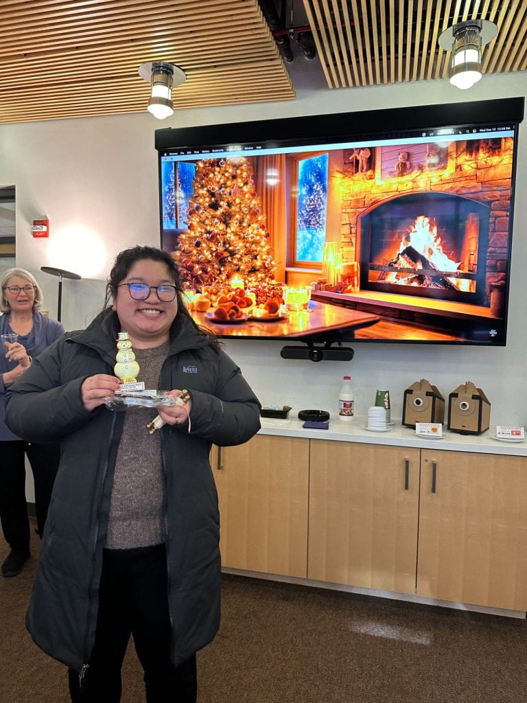 Woman in a jacket and glasses holds a snowman trophy in front of a large monitor displaying a Christmas tree and a roaring fire