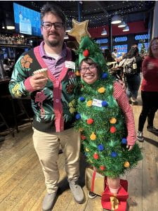 A man in a pink trimmed sweater and a woman wearing a Christmas tree costume.