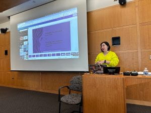Bianca Jimenez, a woman in a bright yellow shirt, stands at the podium in presenting. Her slides are on the projector in the background.