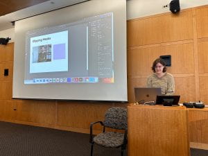 Franny McNamara presents. She stands at the podium in a brown shirt looking intently at her computer. Her screen is sharing on the projector and the audience is viewing her adobe illustrator.