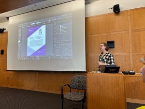 Laura Nevins stands at the podium in a black and white plaid shirt. She stands speaking to the audience with a share of her screen that displays an adobe illustrator flyer on the projector screen in the background. 