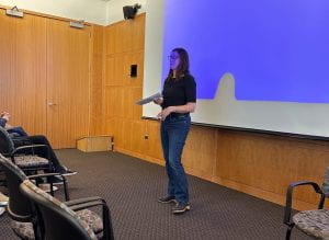 Jill Manor stands in front of a blue projector screen holding a paper handout and addressing the room. 