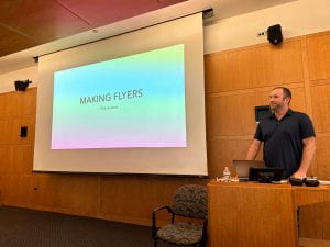 Phil Hoskins, a man with a black shirt, stands at the podium near a projector with a screen displaying "Making Flyers: Phil Hoskins" with an ironically bland design. 